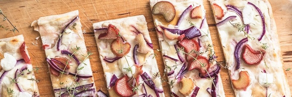 Flammkuchen mit Rhabarber, roten Zwiebeln, Ziegenkäse und Honig liegt in Stücke geschnitten auf einem Holzbrett. 