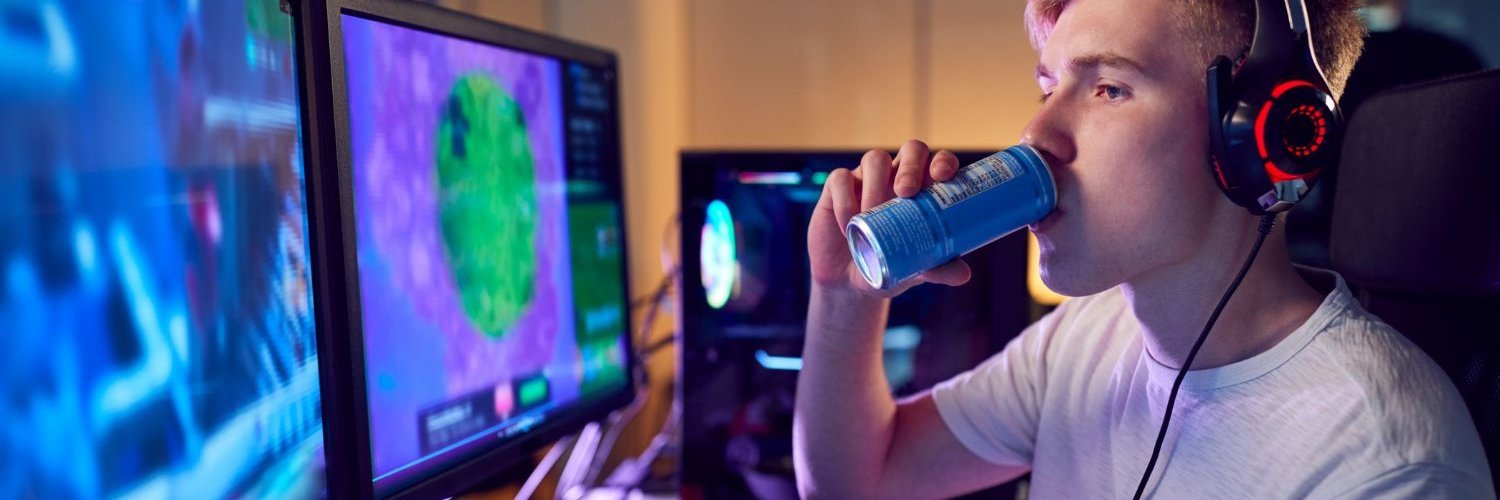 Ein Jugendlicher sitzt mit Kopfhörern vor mehreren Bildschirmen, auf denen man ein Computerspiel sieht, und trinkt aus einer Energydrink-Dose.