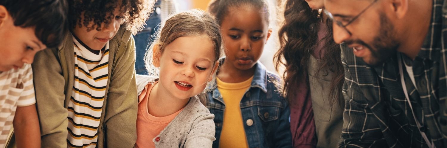 Ein Lehrer und mehrere Grundschulkinder stehen um einen Tisch und beugen sich über verschiedene Materialien.