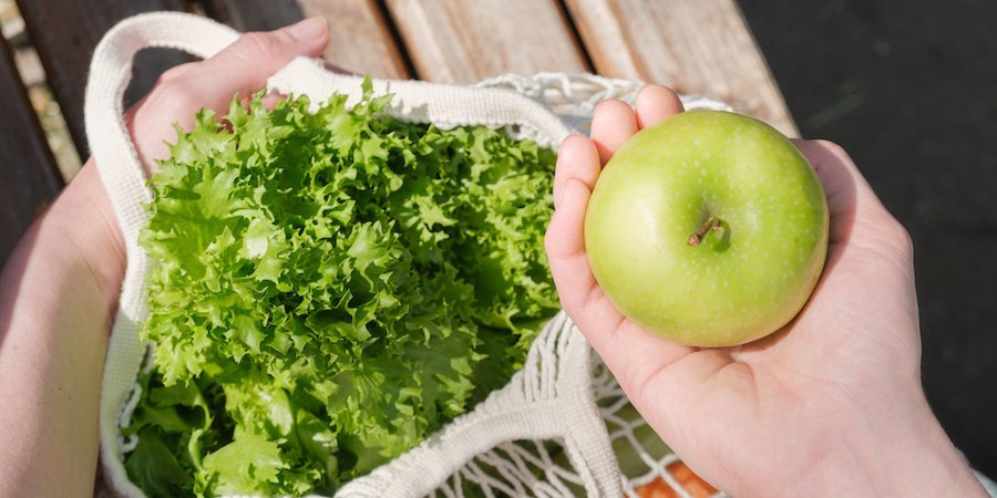 Eine Hand greift in einen Stoffbeutel, in dem sich ein Apfel, ein Salatkopf und anderes Gemüse befindet.