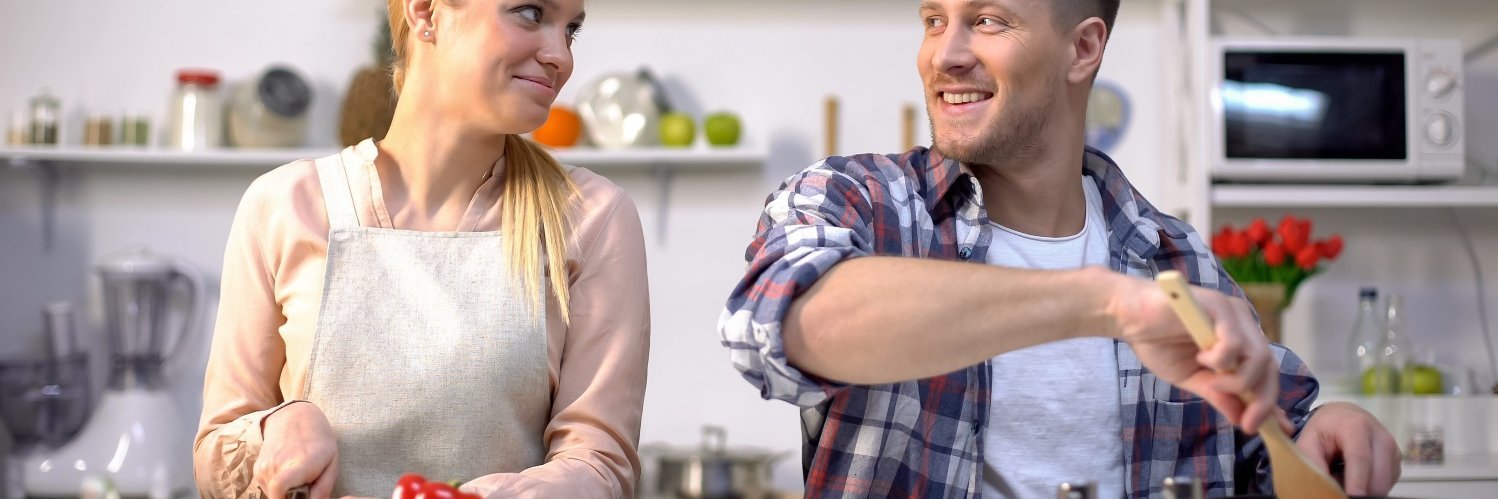 Eine junge blonde Frau und ein dunkelhaariger junger Mann stehen in einer Küche und sehen sich an. Die Frau schneidet Paprika klein und der Mann rührt mit einem Holzlöffel in einer Pfanne auf dem Herd. 