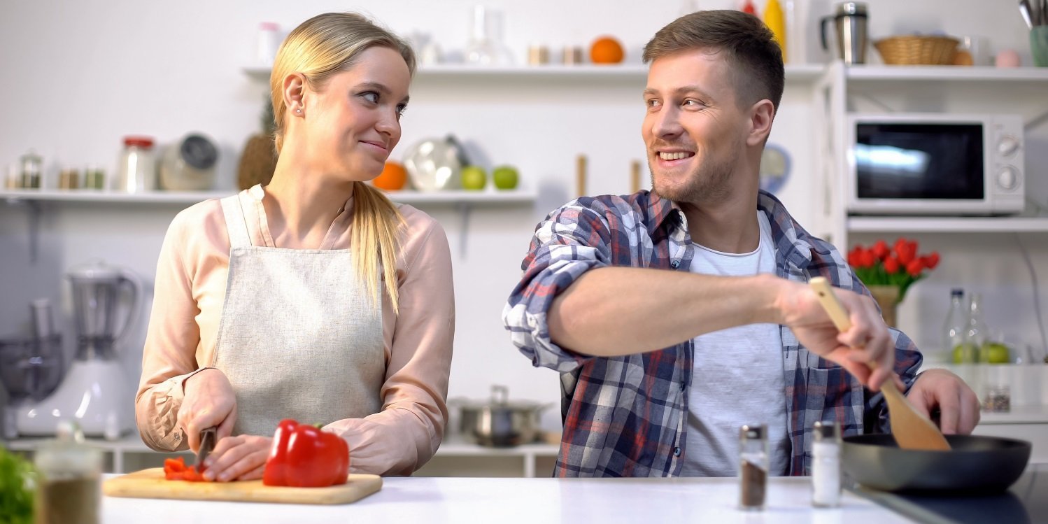 Eine junge blonde Frau und ein dunkelhaariger junger Mann stehen in einer Küche und sehen sich an. Die Frau schneidet Paprika klein und der Mann rührt mit einem Holzlöffel in einer Pfanne auf dem Herd. . Artikel "Kochen ohne Krempel" öffnen.