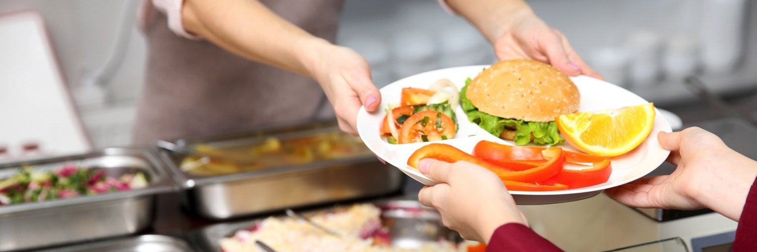 Eine Frau im roten Mantel nimmt an einer Essensausgabe von einer Frau in Schürze einen Teller entgegen, auf dem ein Brötchen, Rohkost, Salat und Obst liegen. 