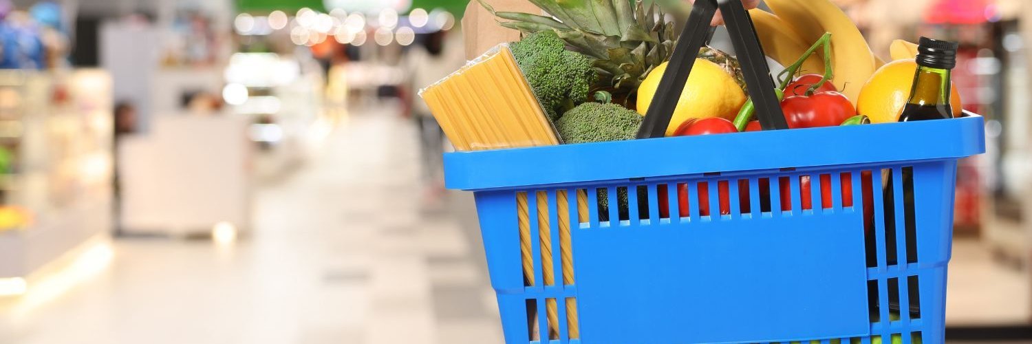 Eine Person im Supermarkt hält einen blauen Einkaufskorb mit Obst, Gemüse, Nudeln und Öl. 
