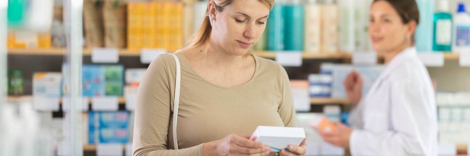 Eine Schwangere steht in der Apotheke und schaut sich eine Verpackung an. 
