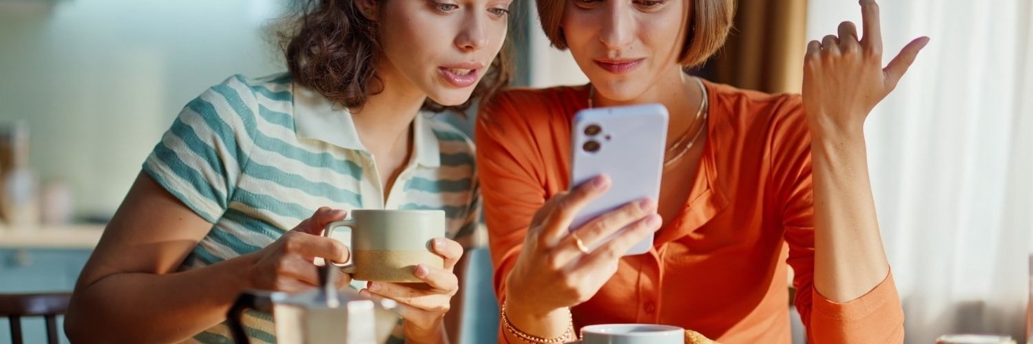Zwei junge Frauen sitzen an einem gedeckten Frühstückstisch und schauen gemeinsam auf ein Handy.