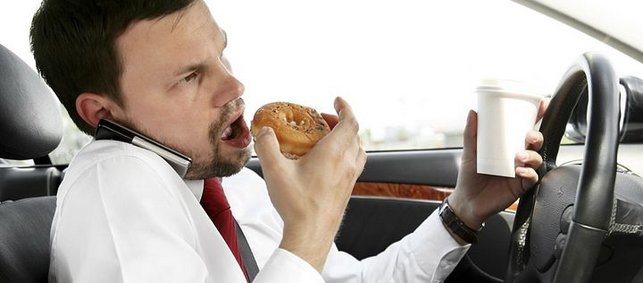 Ein Mann am Steuer eines Autos, der am Essen und Trinken ist und das Handy zwischen Ohr und Schulter eingeklemmt hat