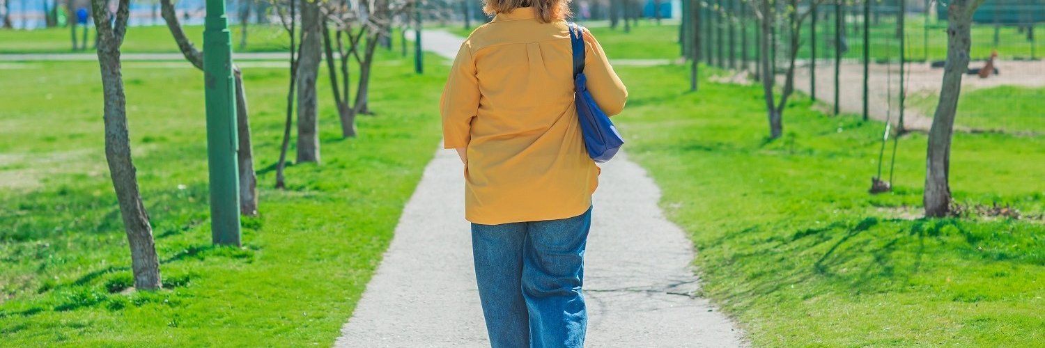 Eine Frau mittleren Alters in gelber Bluse und Jeans läuft eine Allee mit kahlen Bäumen entlang.