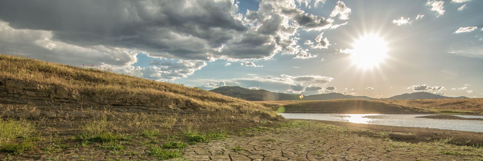 Eine ausgetrocknete Landschaft. Im Hintergrund ein See mit niedrigem Wasserstand. Und darüber scheint eine gleißende Sonne.