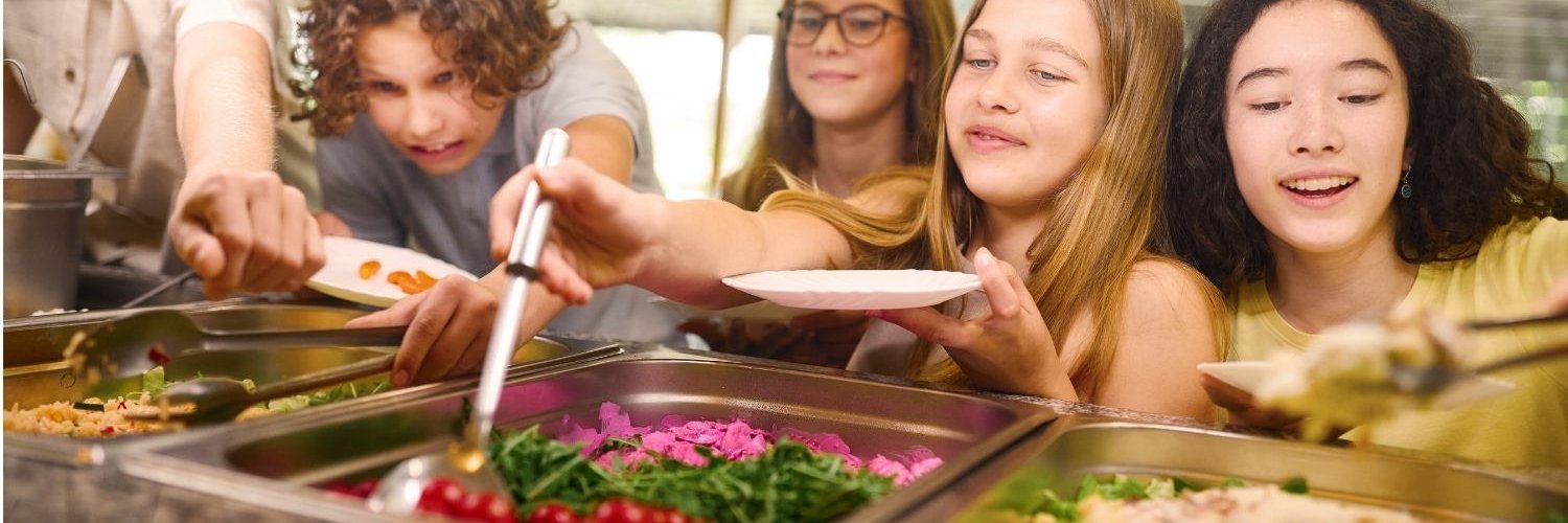 Eine Gruppe von Schülerinnen und Schülern lehnt sich über eine Essensausgabe und bedient sich mit Mittagessen.
