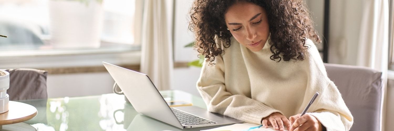 Eine junge Frau sitzt zuhause am Tisch vor ihrem Laptop und macht sich daneben Notizen in ein Heft. 