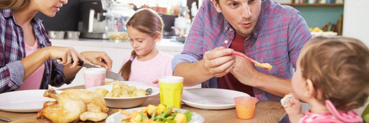 Ein Elternpaar sitzt in der Küche mit zwei Kindern beim Essen. Der Vater füttert das Kleinkind und die Mutter schneidet dem größeren Kind das Essen klein.