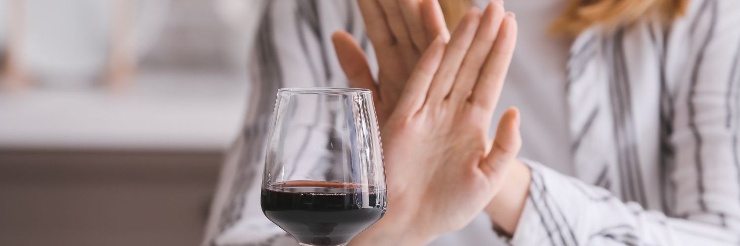 Auf einem Tisch steht ein Glas Rotwein. Eine Frau sitzt davor und macht mit den Händen eine abwehrende Geste.