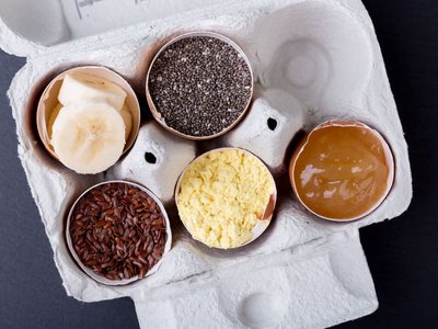 Es ist eine offene 6er-Eierpackung mit Eierschalen zu sehen. In den Eierschalen sind die veganen Alternativen zu Eiern zu sehen: Banane, Chiasamen, Leinsamen, Apfelmus und Ei-Ersatzpulver.