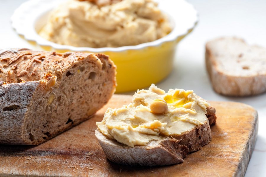 Eine Scheibe Brot mit Hummus. Daneben ein Brot und dahinter eine Schüssel mit Hummus.