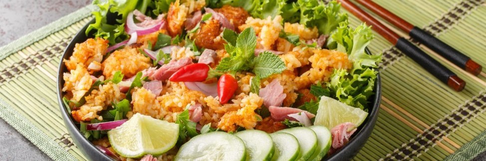 Auf einer Bastmatte steht ein dunkler Teller mit Salatblättern, knusprigem Reis, Gurkenscheiben, Limettenstücken, Chilischoten und Minzeblättern. Daneben liegt ein Paar Essstäbchen.