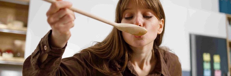 Eine Frau mit langen braunen Haaren und brauner Blus beugt sich über einen Kochtopf und probiert von einem Kochlöffel.
