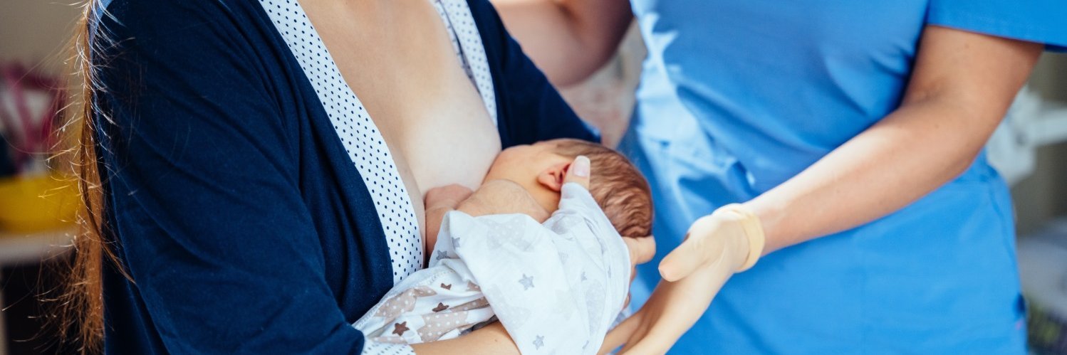 Eine Frau hat ihren Säugling zum Stillen angelegt und eine Person in Krankenhauskleidung steht daneben und legt unterstützend die Hand unter den Kopf des Babys.