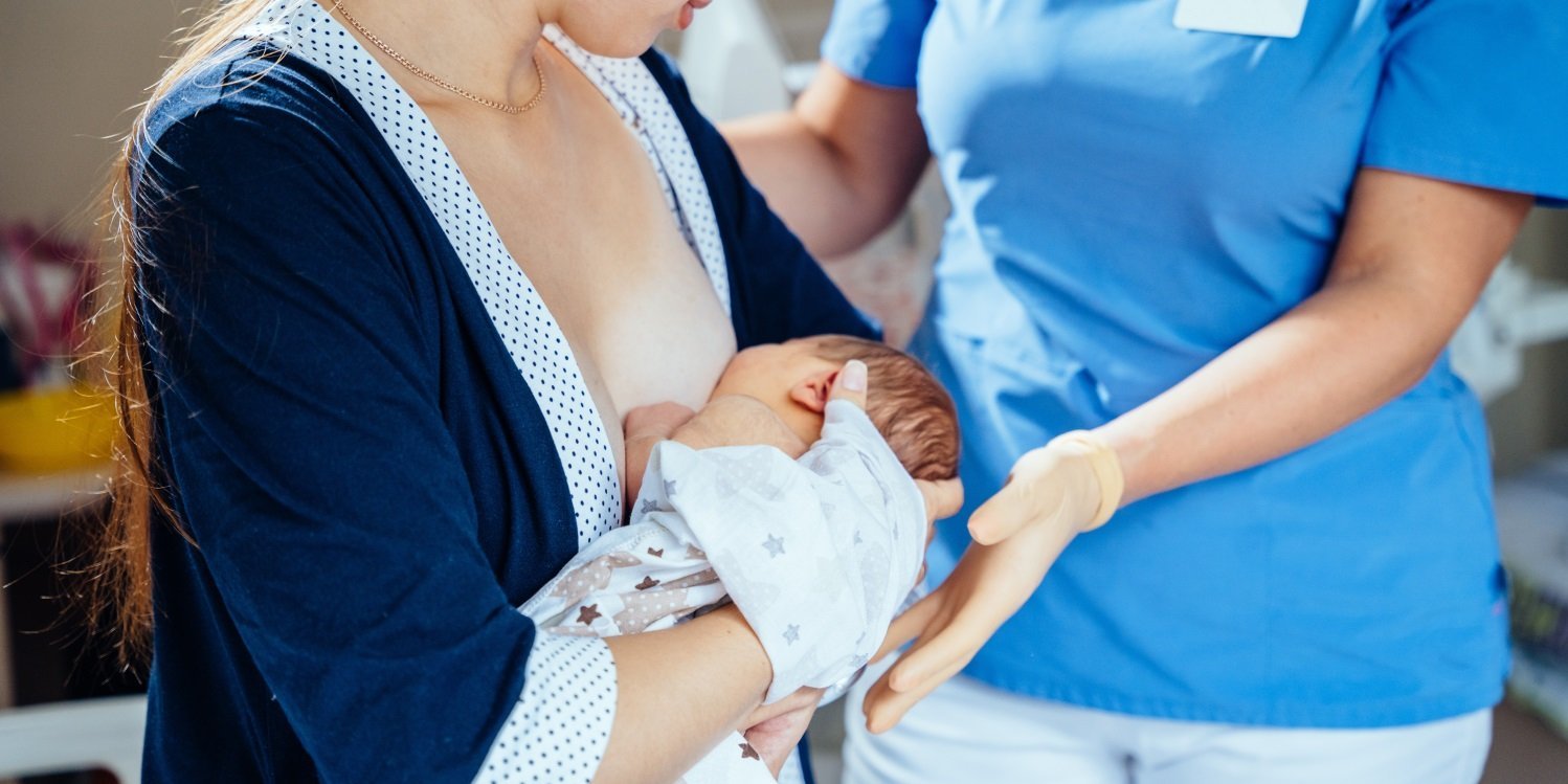 Eine Frau hat ihren Säugling zum Stillen angelegt und eine Person in Krankenhauskleidung steht daneben und legt unterstützend die Hand unter den Kopf des Babys.. Artikel "Schon in der Schwangerschaft ans Stillen denken" öffnen.