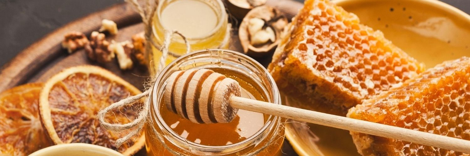 Zwei Gläser Honig auf einem Tablett, daneben steht eine Schale mit Honigwaben und eine mit Pollen. 