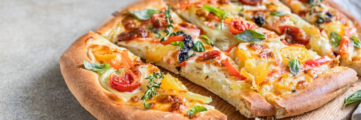 Auf einem Holzbrett liegt eine ovale Pinsa mit verschiedenfarbigen Tomatenstücken, Zwiebeln, Käse und Kräutern. 