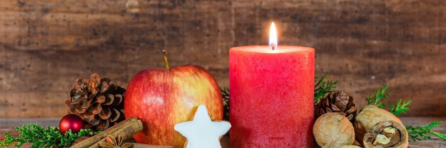 Vor einer Holzverkleidung ist eine brennende rote Kerze dekoriert mit Zweigen, Zapfen, Nüssen und Keksen. 