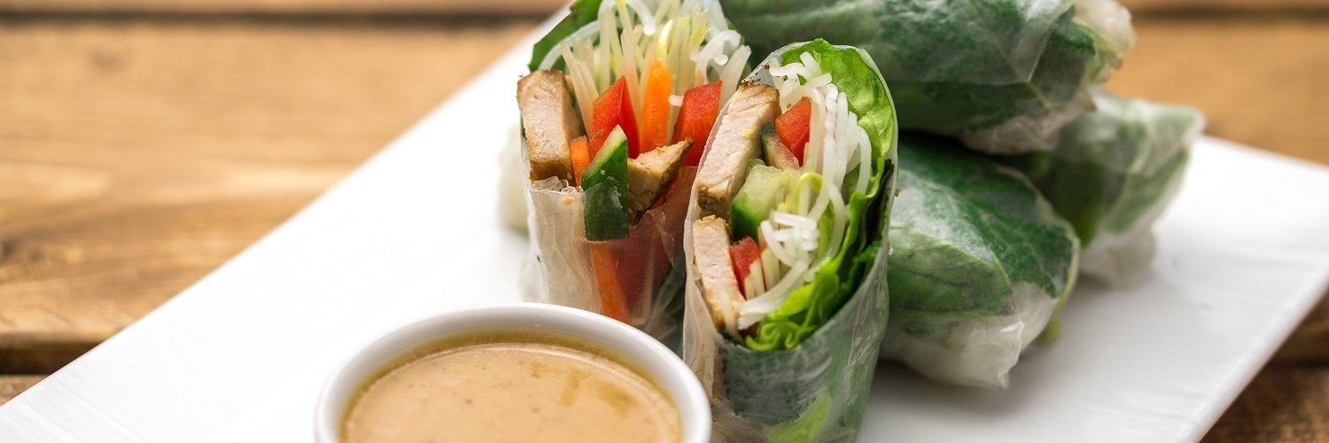 Auf einem weißen Teller liegen Sommerrollen aus Reispapier gefüllt mit Tofu, Paprika, Gurke, Möhren, Salat und Reisnudeln mit einer Schale Erdnusssauce. 