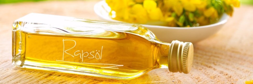 Auf einem Holztisch liegt eine Glasflasche mit gelbem Rapsöl. Im Hintergrund steht eine weiße Schale mit Rapsblüten.