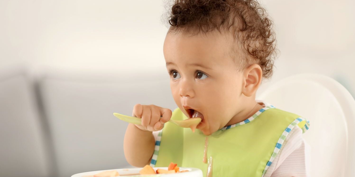 Ein Kleinkind isst mit einem Löffel von seinem Teller.. Artikel " Essen für Kleinkinder" öffnen.
