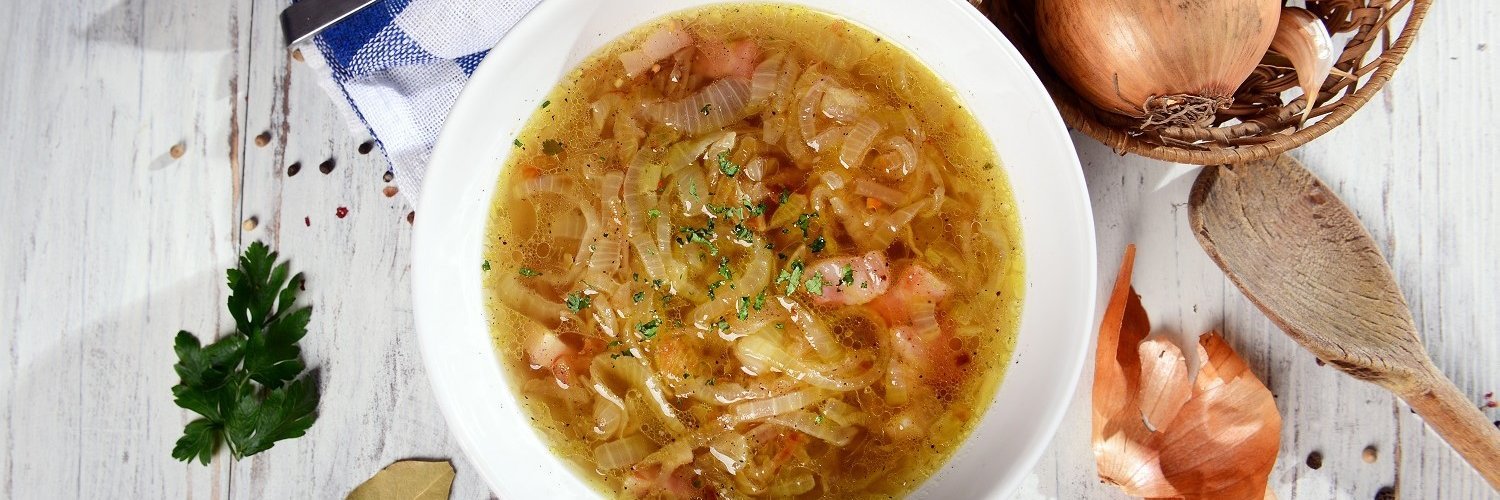 Selbstgemachte Zwiebelsuppe ist in einem weißen Teller angerichtet. Um den Teller herum liegen Petersilie, ein Lorbeerblatt, ein Kochlöffel und ein Bastkörbchen mit Zwiebeln. 