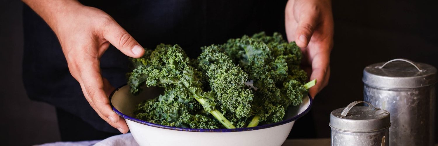Männerhände stellen eine weiße Schüssel mit Grünkohl auf einen Tisch neben ein Geschirrtuch und zwei Dosen.
