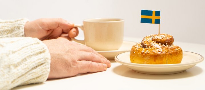 Jemand sitzt am Tisch mit einer Tasse Kaffee und auf einem Teller eine Zimtschnecke mit einer schwedischen Fahne