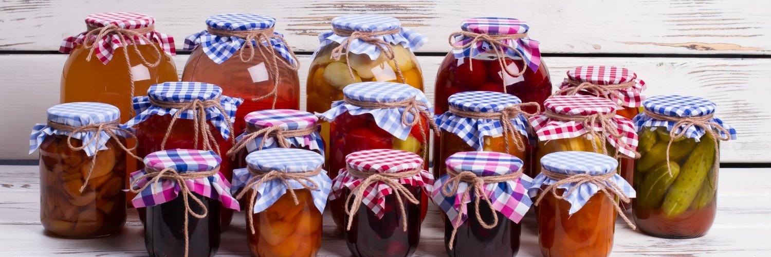 Vor einer hellen Holztäfelung stehen viele verschieden große Einmachgläser mit Obst und Gemüse, dekoriert mit farbig karierten Stoffdeckeln und Schleifen. 
