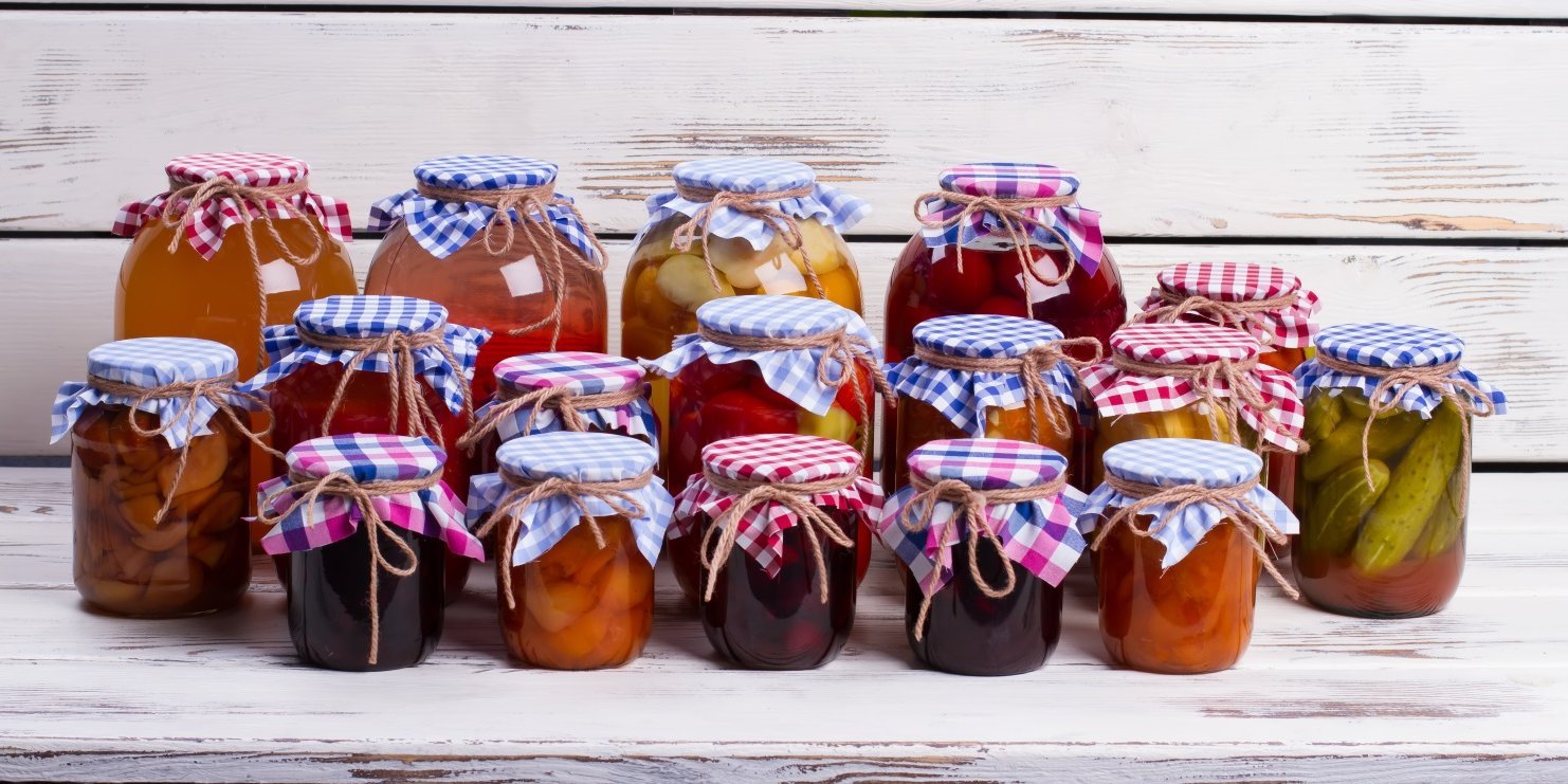 Vor einer hellen Holztäfelung stehen viele verschieden große Einmachgläser mit Obst und Gemüse, dekoriert mit farbig karierten Stoffdeckeln und Schleifen. . Artikel "Den Sommer im Glas verlängern" öffnen.