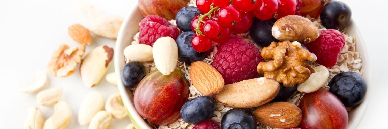 In einer weißen Schale sind Stachelbeeren, Himbeeren, Blaubeeren und Johannisbeeren mit Walnüssen, Mandeln und Paranüssen als Topping auf Haferflocken. 