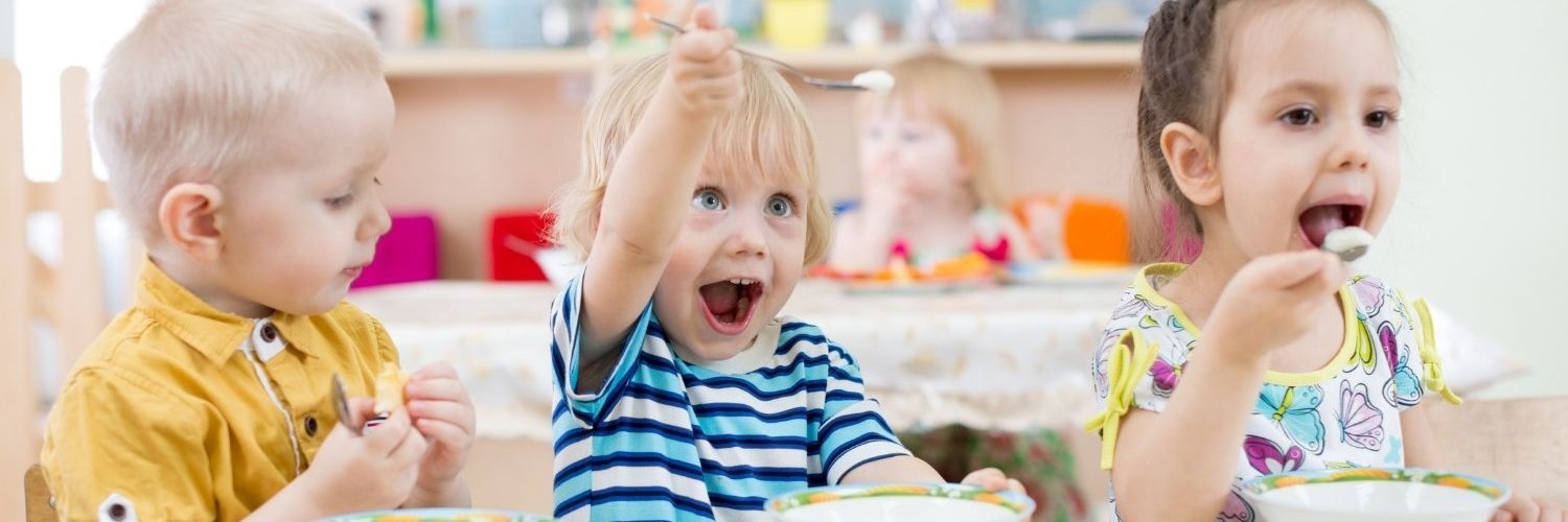 Drei Kindergartenkinder sitzen am Tisch beim Essen. Eins beißt in sein Brot, eins hebt den Löffel in die Luft und eins schiebt sich Essen in den Mund.