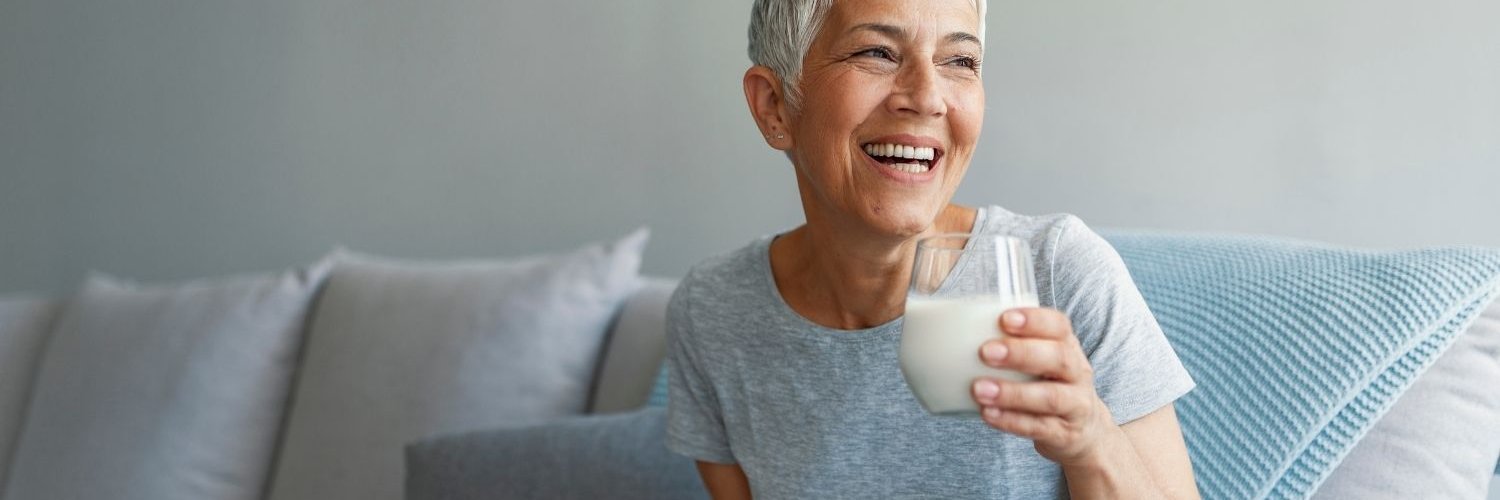 Eine Frau mit kurzen grauen Haaren sitzt lachend auf einem Sofa und hält ein Glas Milch in der Hand. 