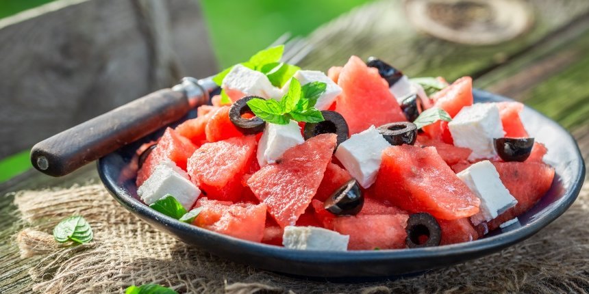 Auf einem Holztisch im Freien steht ein dunkler Teller mit gewürfelter Wassermelone, Fetastücken, schwarzen Oliven und Minzblättern. . Artikel "Sommerküche mit Feta-Käse" öffnen.
