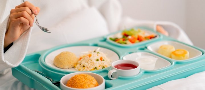 Es ist ein Tablett mit Essen abgebildet, eine Person im Bademantel und einer Gabel in der Hand isst davon. Die Mahlzeit besteht aus einem bunten Salat, Gemüsereis, einem Sesambrötchen und rotem Tee. 