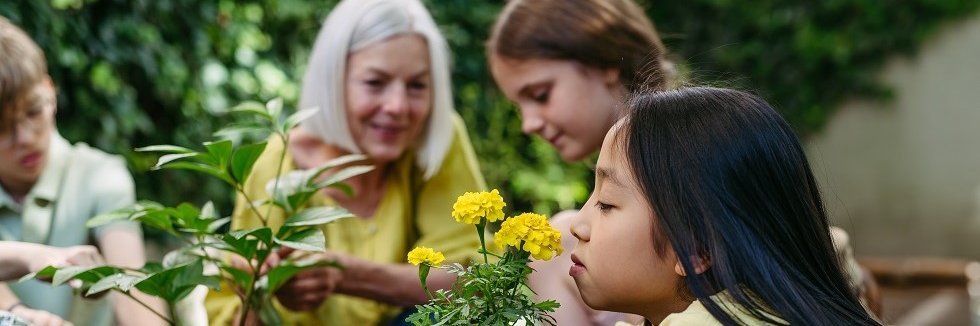 Mehrere Kinder sind mit ihrer Lehrerin im Schulgarten. Im Vordergrund kniet in Mädchen, das mit geschlossenen Augen an einer Blume mit gelben Blüten riecht.  