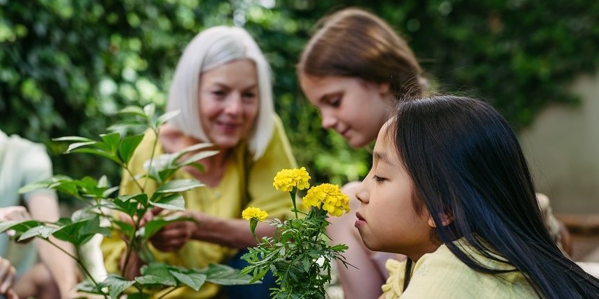 Mehrere Kinder sind mit ihrer Lehrerin im Schulgarten. Im Vordergrund kniet in Mädchen, das mit geschlossenen Augen an einer Blume mit gelben Blüten riecht.  . Artikel "Ernährungsbildung in und außerhalb der Schule" öffnen.