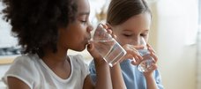 Zwei Mädchen im Grundschulalter trinken jeweils Wasser aus einem Glas. 
