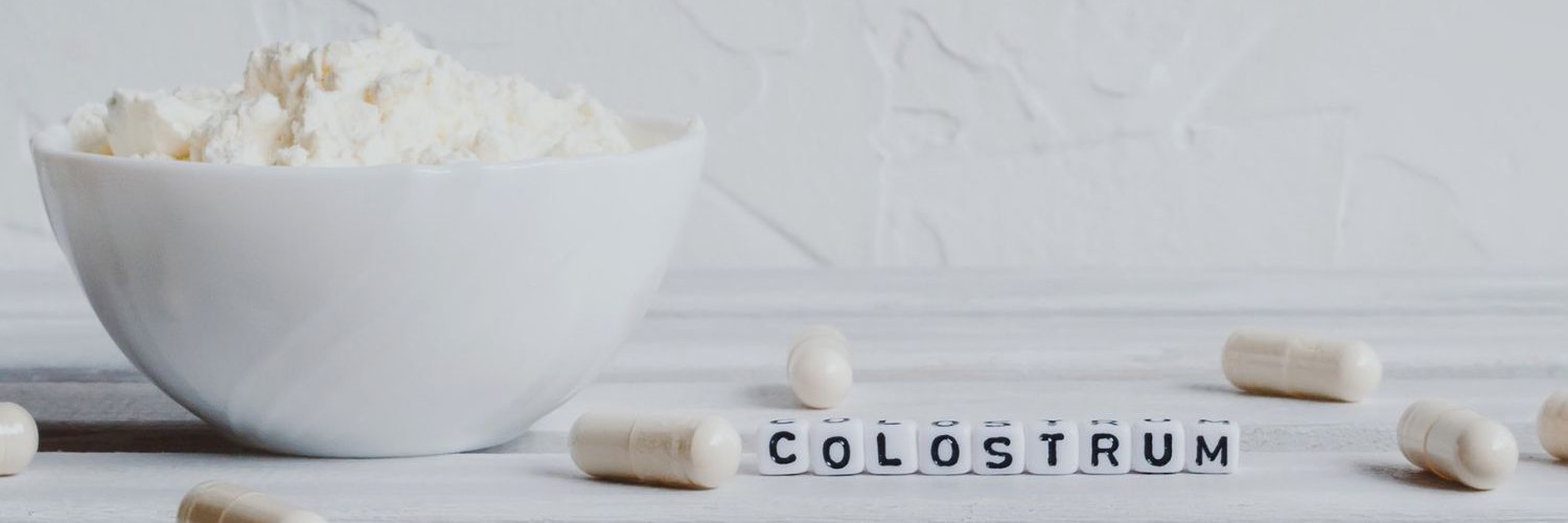 Auf einem weißen Tisch steht eine weiße Schüssel mit Pulver, daneben liegen weiße Kapseln und Buchstabenwürfel die das Wort Colostrum bilden.