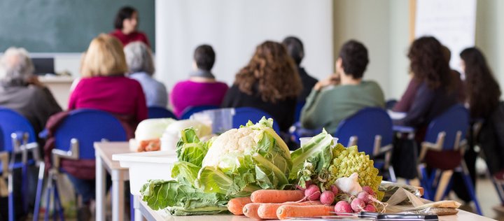 Erwachsene in einem Klassenraum, im Vordergrund steht ein Gemüsekorb auf dem Tisch