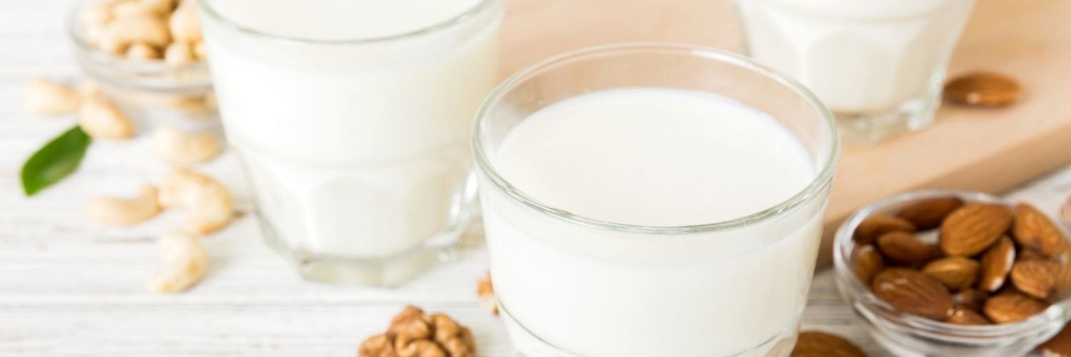 Auf einem hellen Untergrund stehe drei Gläser mit Milchalternativen gefüllt. Daneben liegen jeweils Mandeln, Walnusshälften und Cashews.
