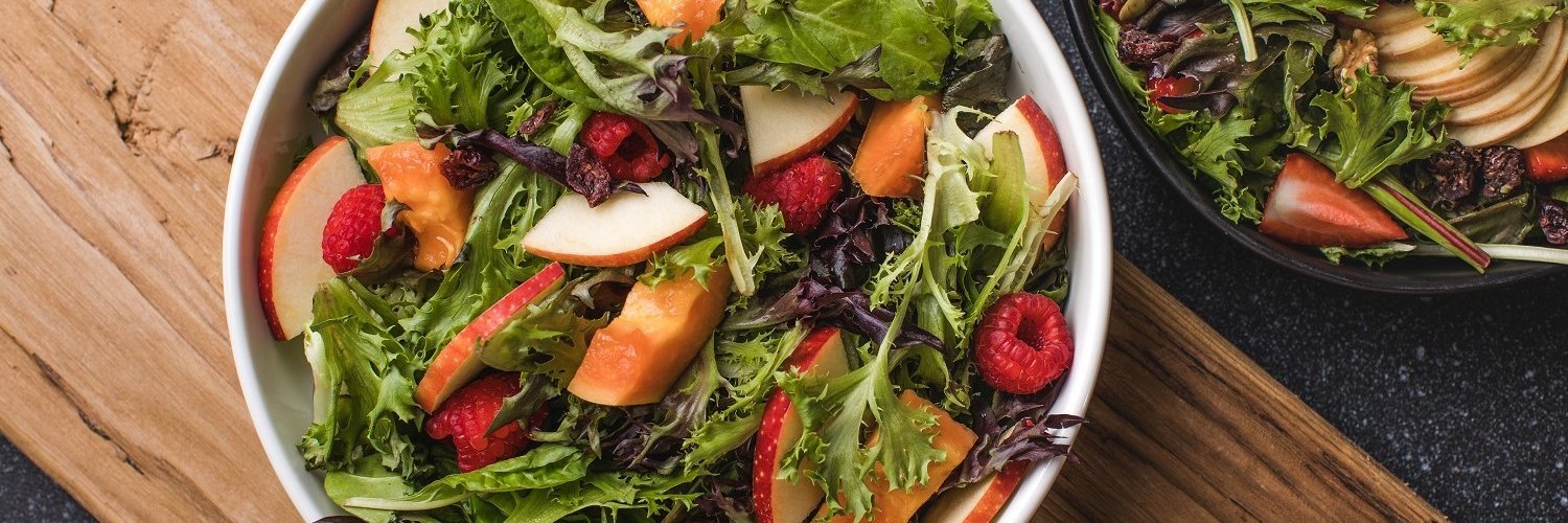 Auf einem weißen Teller ist ein Salat aus Rucola, Apfelstücken, Himbeeren und gebackenen Käsenuggets angerichtet.