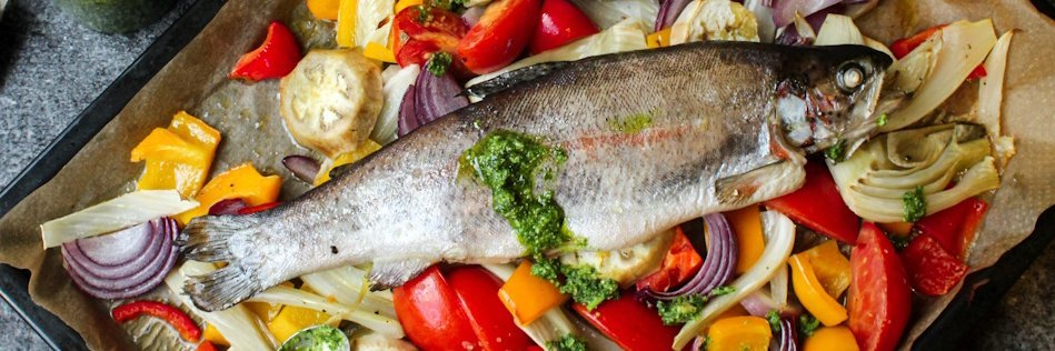 Auf einem Backblech mit buntem Gemüse liegt ein ganzer Fisch.