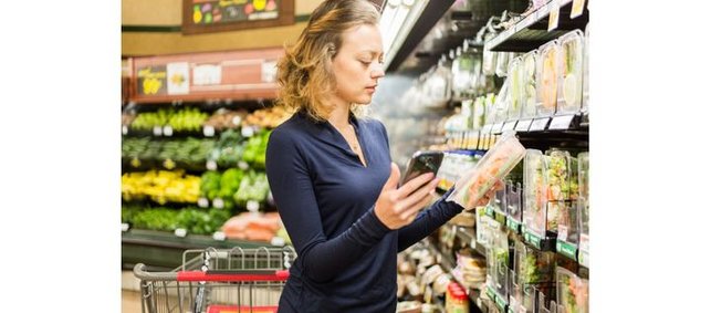 eine Frau steht vor einem Supermarktregal und hat ein Produkt sowie ihr Handy in der Hand