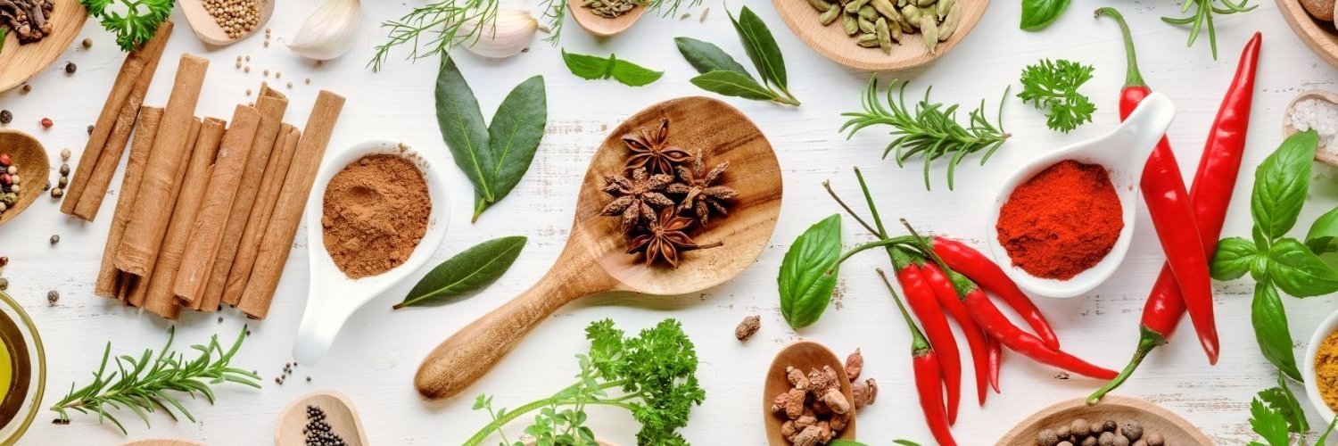 Auf einer weißen Fläche liegen verstreut frische Lorbeerblätter, Rosmarin, Basilikum, Dill und Petersilie. Außerdem Holzlöffel und Behälter mit Senfkörnern, Paprikapulver, Zimtstangen, Kardamom, Sternanis, Muskatnüssen, Peperoni und Knoblauchzehen.