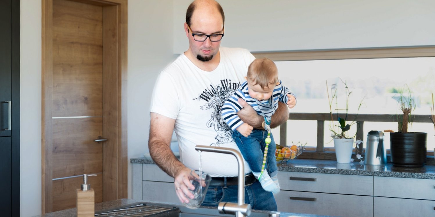 Ein Mann hat einen Baby auf dem Arm und lässt Wasser aus einem Hahn in ein Glas laufen.. Artikel "Wasser: Gesundheit und Umwelt" öffnen.